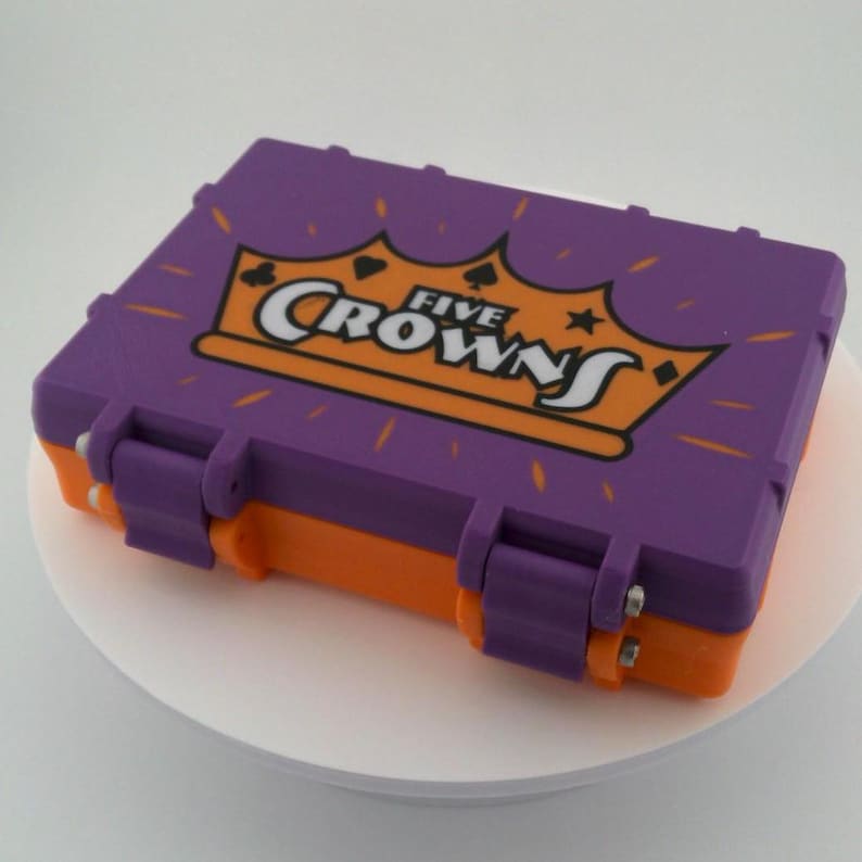 May include: A purple and orange rectangular case with the words "FIVE CROWNS" in white and orange. The case has a crown graphic and is closed with two hinges.