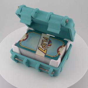 May include: An open, teal-colored case holding a deck of cards. The cards are labeled "FLIP 7" and have a teal, yellow, and red design. The case has a hinged lid and metal clasps.