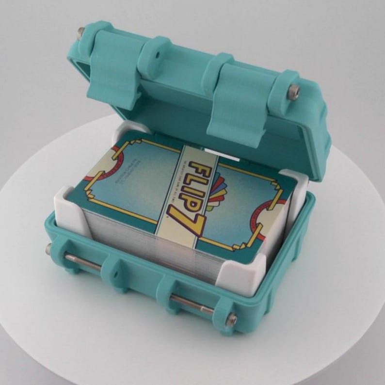 May include: An open, teal-colored case holding a deck of cards. The cards are labeled "FLIP 7" and have a teal, yellow, and red design. The case has a hinged lid and metal clasps.