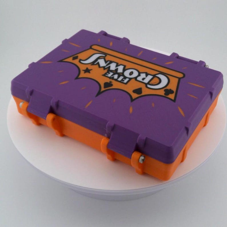 May include: A purple and orange plastic case with the words "FIVE CROWNS" in white lettering. The case has a rectangular shape with a hinged lid and clasps. The case is sitting on a white surface.