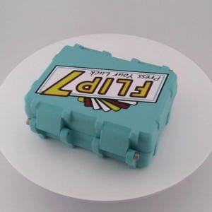 May include: A teal-colored rectangular box with the text "FLIP 7 Press Your Luck" in yellow and white. The box has a hinged lid and metal clasps. The design includes a stylized graphic with red, yellow, and white elements.