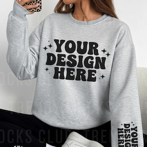 May include: A light gray sweatshirt with the text "YOUR DESIGN HERE" in black, accented with stars. The design is repeated on the sleeve. The sweatshirt is part of a mock-up, with the Trendy Mocks Club logo visible.