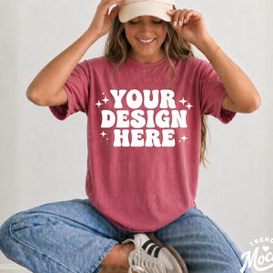 May include: A maroon t-shirt with the text "YOUR DESIGN HERE" in white, accented with starbursts. The person is wearing a beige baseball cap and blue jeans. The shirt is a mock-up for a design.