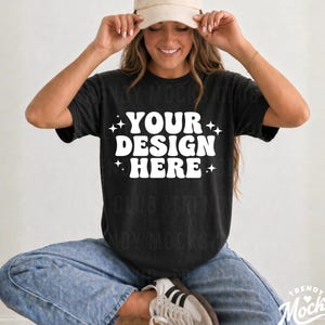 May include: Black t-shirt with the text "YOUR DESIGN HERE" in white. The model is wearing a beige baseball cap and blue jeans. The shirt is a crew neck and has short sleeves.