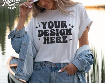 May include: White t-shirt with the text "YOUR DESIGN HERE" in a black, retro font. The model is wearing a white baseball cap and a light blue denim jacket. The shirt is paired with light blue jeans.