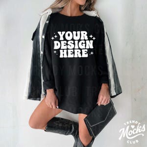 May include: Black long-sleeve sweatshirt with the text "YOUR DESIGN HERE" in white. The model is wearing a black leather jacket with a white fur lining, black boots, and holding a black clutch. The image is a mockup for apparel.