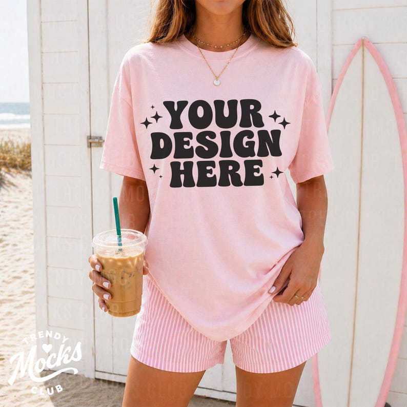 May include: A light pink t-shirt with the text "YOUR DESIGN HERE" in black, paired with pink and white striped shorts. The person is holding a clear plastic cup with iced coffee. A surfboard is in the background.