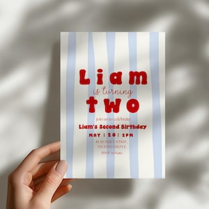 Puede incluir: Una invitación de cumpleaños con un fondo a rayas azul claro y blanco. El texto "Liam is turning two" es rojo, con detalles de la fiesta en letra roja más pequeña. La invitación es sostenida por una mano.