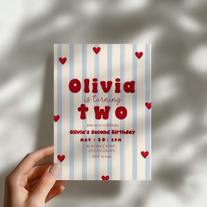 May include: A rectangular birthday invitation card featuring a light blue and white vertical striped background with scattered small red hearts. The text reads: Olivia is turning two, join us to celebrate, Olivia's Second Birthday, May 20, 2PM, 81 Sunset Strip, Ocean Grove, RSVP to Kate.