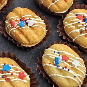 May include: Several golden-brown oval-shaped baked goods in brown paper liners, each decorated with white icing drizzles, small red and blue spherical sprinkles, and red, white, and blue star-shaped sugar decorations arranged on top.