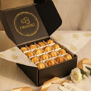 May include: A black cardboard box containing sixteen nut-shaped cookies with caramel filling, arranged in a grid. The lid features a gold circular logo with the text ORESHKI. Two cookies sit outside the box, and a tag reads For You.