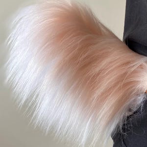 Peut inclure: Accessoire en fausse fourrure duveteuse avec un dégradé de blanc à rose clair. La fourrure est longue et texturée, créant un aspect doux et volumineux. L'article est probablement un élément de costume ou décoratif.