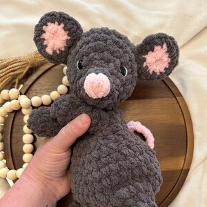 May include: A handmade gray crochet mouse plush toy with pink accents on the ears and nose. The stuffed animal has black safety eyes and a pink tail. The toy is designed to be soft and cuddly.