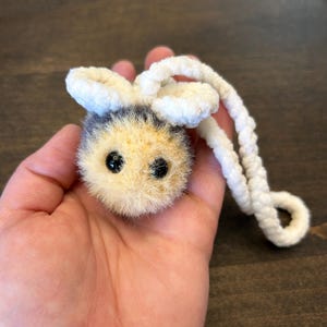 May include: A handmade plush bee with a fuzzy body in shades of yellow, black, and cream. It has large, black eyes and white, fluffy antennae. A white cord is attached for hanging.