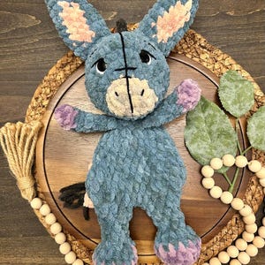 May include: A plush donkey toy crafted from soft blue yarn, featuring long ears with light pink inner accents, a cream-colored muzzle, and purple-tipped hooves. The stuffed animal has embroidered black eyes and a small black tail.