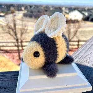 May include: A crocheted bee plush toy with gray and yellow stripes and white wings. The bee is sitting on a white post with a blurred background of houses and a blue sky.