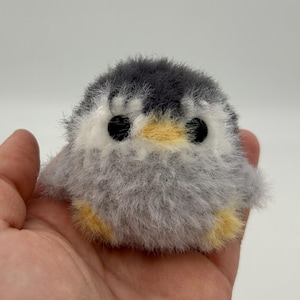 Puede incluir: Un pequeño pingüino de peluche gris y blanco con ojos negros y un pico amarillo. El pingüino está sentado en una mano.