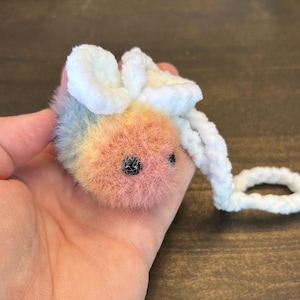 May include: A small, fuzzy, rainbow-colored bee plush toy with white wings and a white cord.