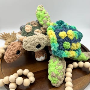 May include: Two handmade crochet sea turtles in shades of brown, green, and blue. The turtles have black safety eyes and are made with a soft, textured yarn. One turtle is light brown with green and gray spots, and the other is blue, green, and yellow.