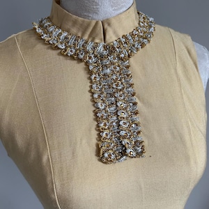 May include: A vintage-style, tan-colored dress with a high collar and a decorative collar necklace. The necklace features a woven design with silver and gold metallic threads, creating a textured and elegant look. The dress is displayed on a mannequin.