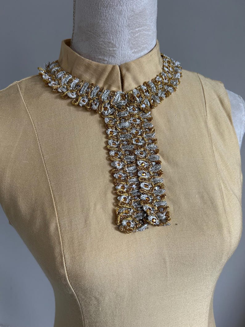 May include: A vintage-style, tan-colored dress with a high collar and a decorative collar necklace. The necklace features a woven design with silver and gold metallic threads, creating a textured and elegant look. The dress is displayed on a mannequin.