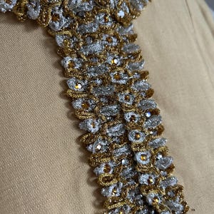 May include: Close-up of a decorative collar with intricate gold and silver metallic threadwork. The collar features a repeating pattern of woven elements and small, sparkling embellishments. The background is a solid, light tan fabric.