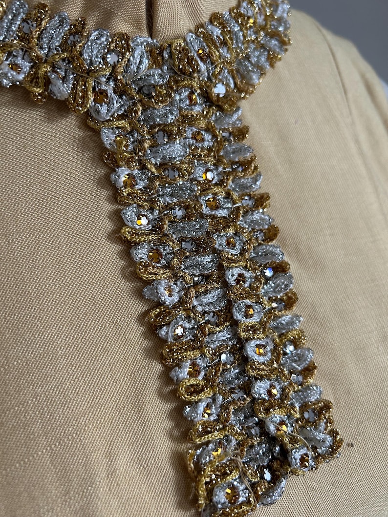 May include: Close-up of a decorative collar with intricate gold and silver metallic threadwork. The collar features a repeating pattern of woven elements and small, sparkling embellishments. The background is a solid, light tan fabric.