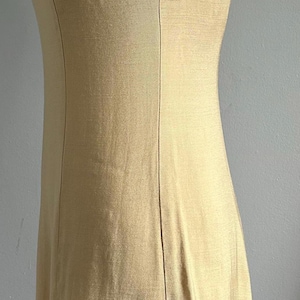 May include: A sleeveless, golden-yellow dress with a high neckline. The dress features a decorative beaded trim around the neckline and a tailored fit. Vertical seams run down the front, adding structure to the garment. The dress is displayed on a mannequin.