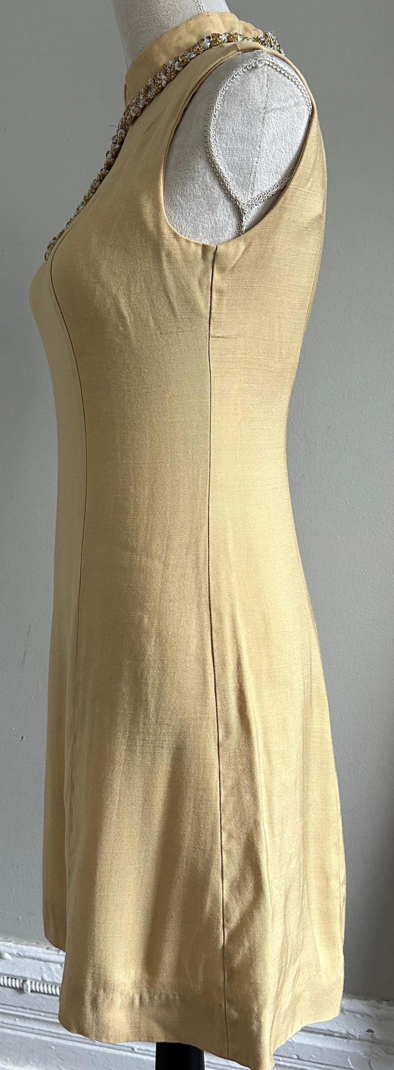 May include: A sleeveless, golden-yellow dress with a high neckline. The dress features a decorative beaded trim around the neckline and a tailored fit. Vertical seams run down the front, adding structure to the garment. The dress is displayed on a mannequin.