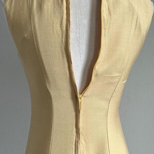 May include: A sleeveless, pale yellow dress with a high neckline and a back zipper. The dress is fitted and appears to be made of a smooth fabric. The neckline is embellished with a decorative trim.