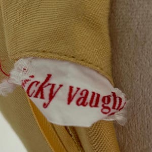 May include: Close-up of a tan garment with a white tag. The tag has the name "icky vaugh" printed in red script. The garment appears to be made of a smooth fabric.