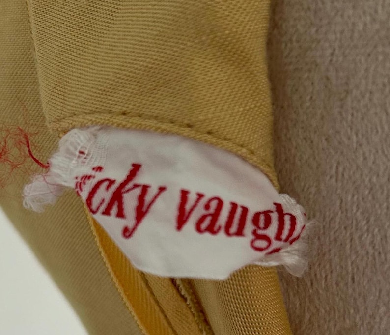 May include: Close-up of a tan garment with a white tag. The tag has the name "icky vaugh" printed in red script. The garment appears to be made of a smooth fabric.