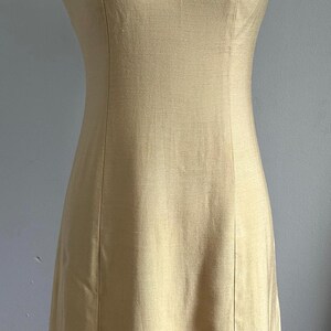 May include: A sleeveless, pale yellow dress with a high neckline and a decorative collar. The collar features a detailed pattern of gold and silver embellishments. The dress has a fitted bodice and a straight skirt.