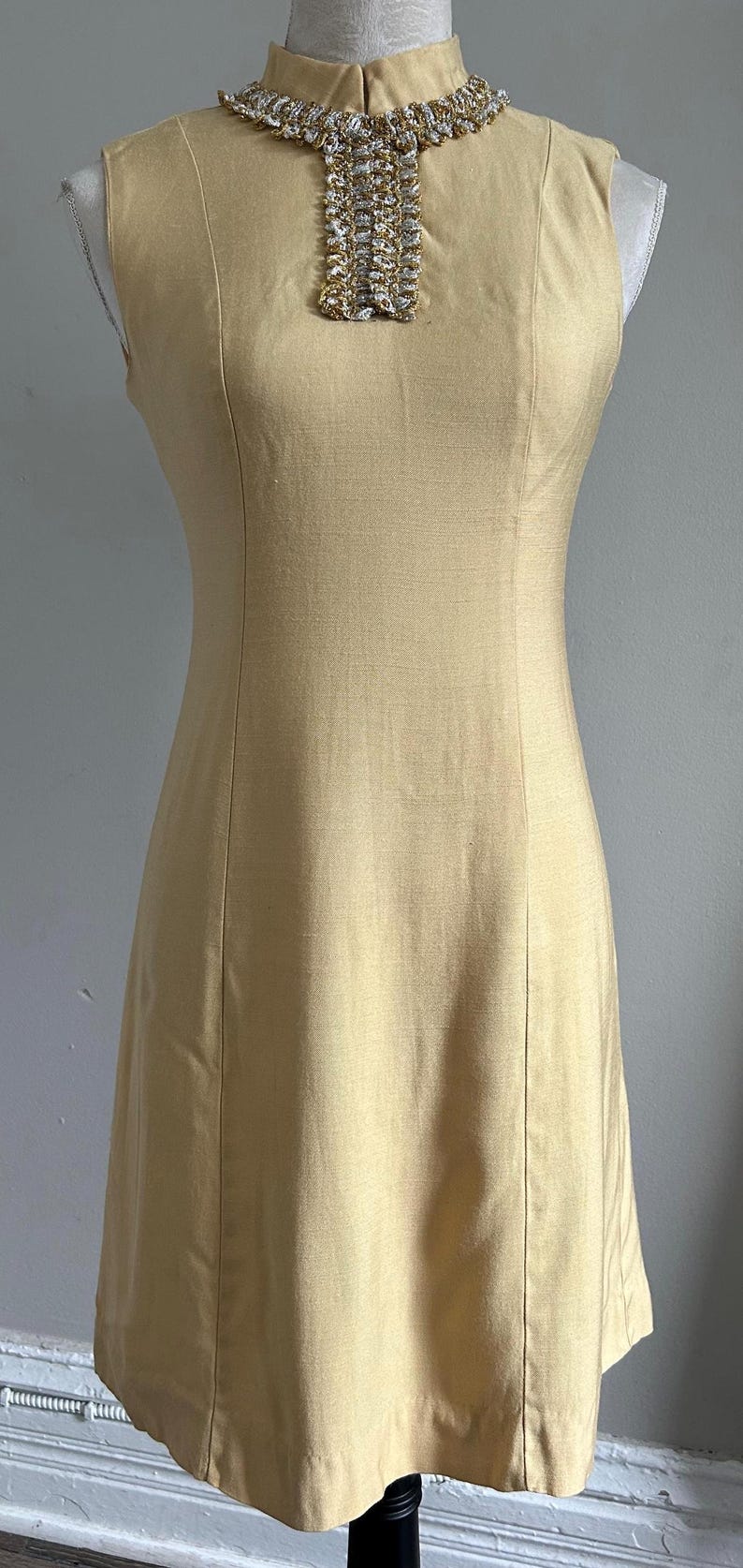 May include: A sleeveless, pale yellow dress with a high neckline and a decorative collar. The collar features a detailed pattern of gold and silver embellishments. The dress has a fitted bodice and a straight skirt.