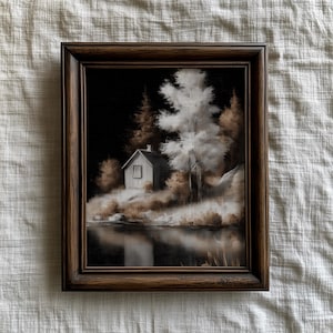 Puede incluir: Una pintura de paisaje enmarcada representa una pequeña cabaña blanca entre árboles junto a un lago. La obra de arte utiliza una paleta de colores apagados de marrones, blancos y negros, creando una estética serena y vintage. El marco es marrón oscuro.