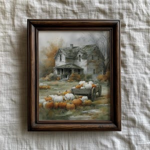 May include: Framed artwork depicting a rural autumn scene. A weathered white house with a green roof is surrounded by pumpkins in shades of orange and white. A wooden cart is filled with pumpkins. The frame is dark brown.