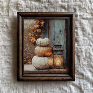 May include: Framed artwork featuring a stack of three pumpkins in shades of white and orange, with a lit lantern and autumn foliage. The artwork is set against a weathered blue wooden backdrop, all within a dark brown frame.