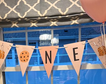 Op de afbeelding: Een roze banner met het woord "ONE" in zwarte letters, versierd met koekjes- en kaarsillustraties. De banner is gespannen tussen roze en gouden ballonnen, perfect voor een eerste verjaardag.