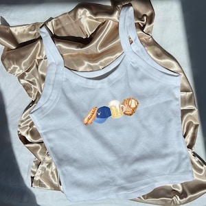 May include: White vest top featuring a sports-themed design. The design includes a hot dog, baseball cap, beer mug, and baseball glove. The top is made of a soft, lightweight material.
