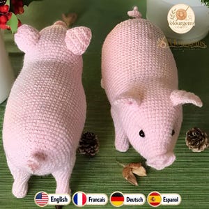 May include: Two handmade, light pink crochet pig figurines. The pigs have small ears, black eyes, and curled tails. The crochet work is detailed, and the pigs are posed on a green surface. The image also includes pinecones and a logo.