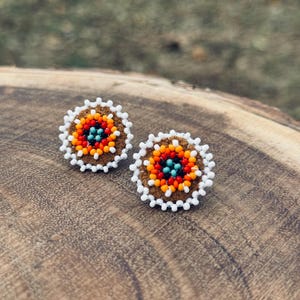 May include: A pair of beaded earrings with a brown suede backing. The earrings are round and feature a colorful geometric pattern in shades of orange, red, white, and blue.