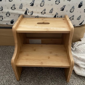 May include: A two-step wooden step stool made of light-colored bamboo. The stool has a rectangular top step with a handle cutout and a lower step. The background features a white sheet with a penguin pattern.