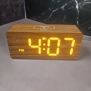 May include: A rectangular wooden digital alarm clock displaying "4:07 PM" in bright yellow digits. The clock has a wood grain finish and a "Snooze" button on top. The brand name "ascece" is also visible.