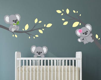 May include: A white crib with a gray wall behind it. There are three koala bear decals on the wall, one with a baby koala, one with a mother koala holding a baby koala, and one with a koala holding a leaf. The decals are on a branch with yellow leaves.