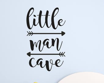 Op de afbeelding: Zwart-witte wanddecoratie met de tekst "little man cave" en twee pijlen op elke regel. De decoratie bevindt zich op een lichtblauwe muur boven een wit bed met een blauw-wit gestreept dekbed.