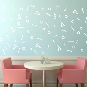 Op de afbeelding: Een lichtblauwe muur met witte geometrische vormen, een witte tafel met een licht houten blad en twee roze stoelen. De tafel is gedekt met een witte servet en een kleine plant.