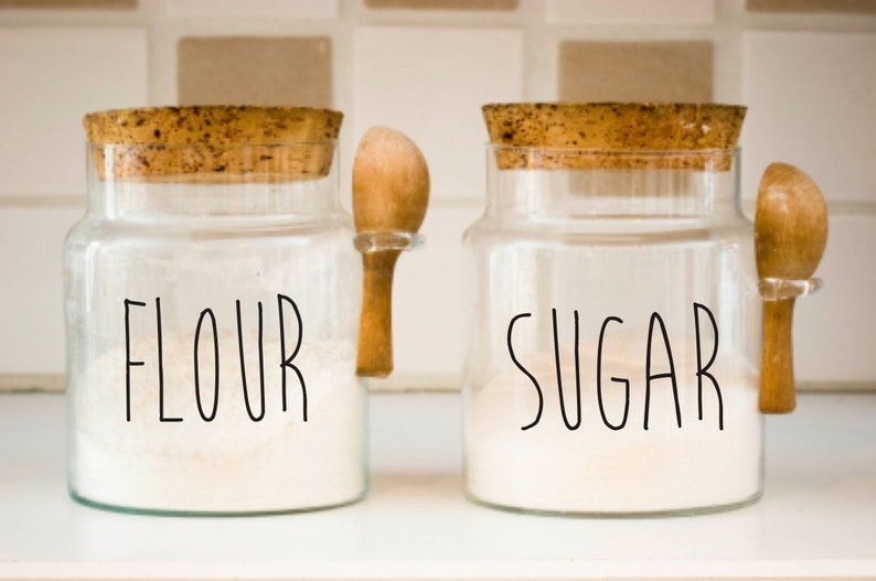 May include: Two glass jars with cork lids and wooden spoons. The jars are filled with white powder and labeled "FLOUR" and "SUGAR".