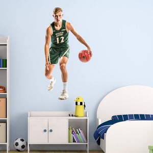 May include: A basketball player wearing a green and white jersey with the text "CAA William & Mary 12" dribbling a basketball. The player is in mid-air, with one foot off the ground. The background is a light blue wall.