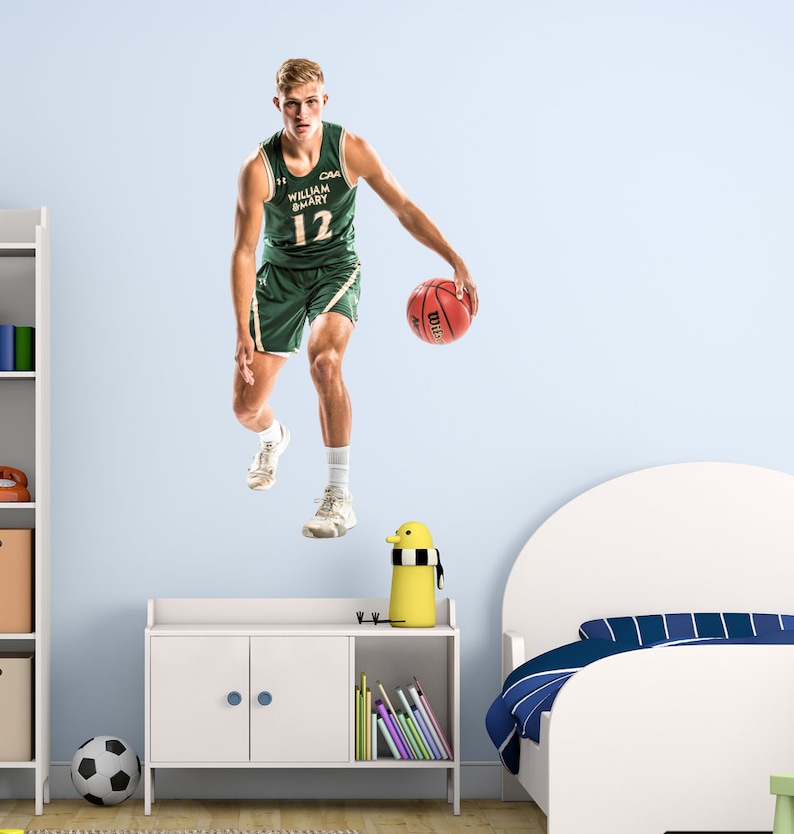 May include: A basketball player wearing a green and white jersey with the text "CAA William & Mary 12" dribbling a basketball. The player is in mid-air, with one foot off the ground. The background is a light blue wall.