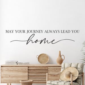Op de afbeelding: Een muursticker met de tekst "May your journey always lead you home" in een cursief lettertype. De sticker is aangebracht op een witte muur in een woonkamer.